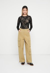 Body lungo trasparente nero a maniche lunghe con motivo floreale abbinato a pantaloni cargo color sabbia con tasche laterali e vestibilità comoda, indossato con scarpe nere.