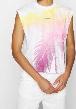 Ærmeløs hvid top med gul og pink palmeprint og "Calvin Klein Jeans"-logo, båret med hvide bukser, hænderne sammenfoldet ved taljen.
