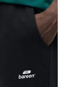 Το χέρι ενός ατόμου ακουμπά μέσα στην τσέπη μαύρου παντελονιού με λευκό λογότυπο «bareen» κοντά στην τσέπη, φοράει πράσινο τοπ.