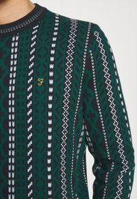 Suéter de punto con patrones geométricos en verde, negro y blanco. Presenta un cuello redondo y un pequeño logo bordado en dorado.