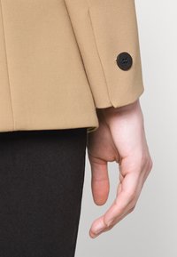Main détendue le long du corps portant un blazer beige avec un bouton noir sur le poignet et un pantalon noir, sur un fond clair uni.