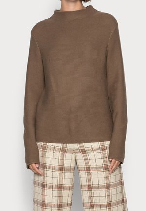 Maglione marrone con texture, col alto e maniche lunghe, abbinato a pantaloni a quadri crema con linee arancioni.