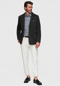 Homme portant un blazer noir, une chemise bleue à motifs, un pantalon blanc, une ceinture noire et des mocassins noirs, debout devant un fond clair uni.