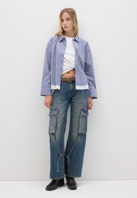 Camicia button-up a righe blu e bianche sopra una maglietta bianca corta, abbinata a jeans cargo a gamba larga e stivali neri alla caviglia.