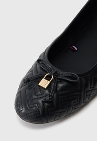 Μαύρη δερμάτινη μπαλαρίνα με υφασμάτινο γεωμετρικό σχέδιο, φιόγκος στη μύτη και μικρή χρυσή πόρπη. Το εσωτερικό είναι μαλακό και σκούρο.