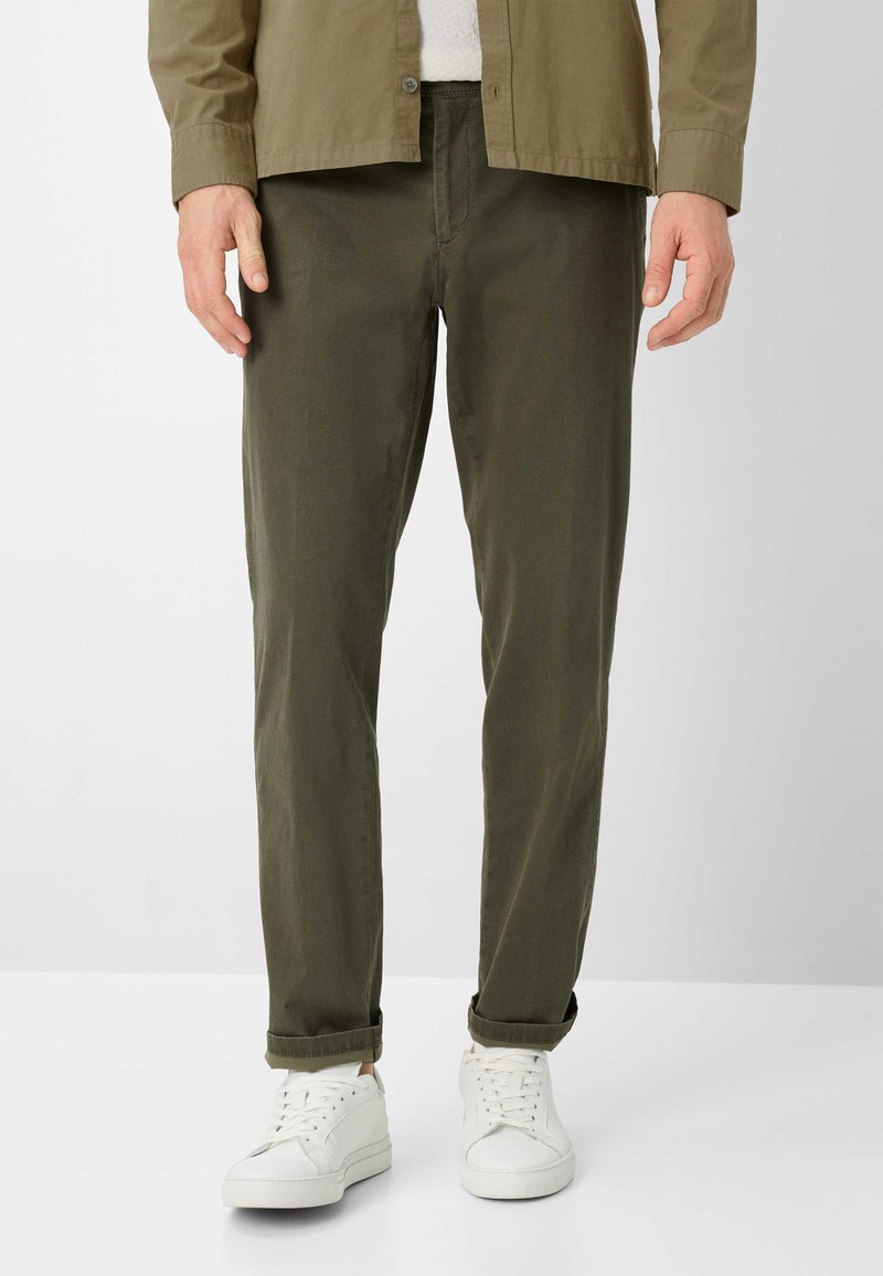 Mann, der olivgrüne Hose, weiße Turnschuhe und ein olivgrünes Hemd trägt, steht vor einem schlichten hellgrauen Hintergrund.