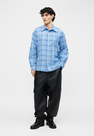 Νέος με σγουρά μαύρα μαλλιά, φορώντας μπλε καρό πουκάμισο με κουμπιά, μαύρο φαρδύ παντελόνι cargo και μαύρα παπούτσια με κορδόνια.