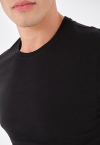 T-shirt in cotone nero con collo a giro e maniche corte. Texture liscia e design aderente, evidenziando un primo piano del colletto e della parte superiore del petto.