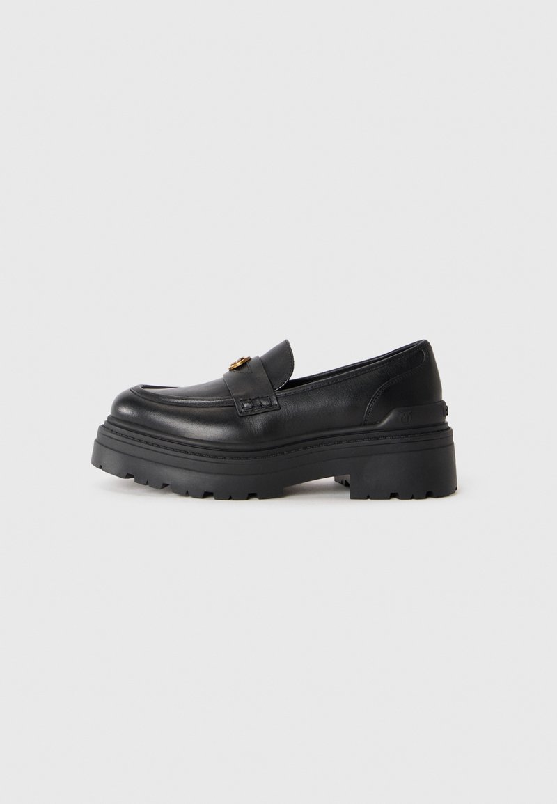 Mocassin en cuir noir avec semelle crantée épaisse et détail de boucle dorée, présenté sur un fond gris clair uni.