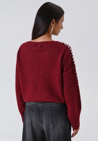 Donna con lunghi capelli scuri che indossa un maglione lavorato a maglia borgogna con dettagli bianchi sulle maniche e jeans neri sbiaditi, di spalle.