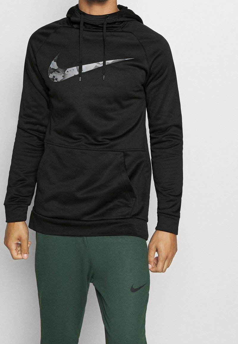 Sudadera negra de Nike con un gran logo de camuflaje, tejido texturizado, bolsillo tipo canguro y capucha con cordón. Combinada con pantalones de chándal verdes de Nike.