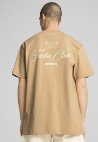 Sandfarvet bomulds-T-shirt med løs pasform. Har et trykt "Turbo Club" logo og grafik på ryggen. Korte ærmer og rund halsudskæring.