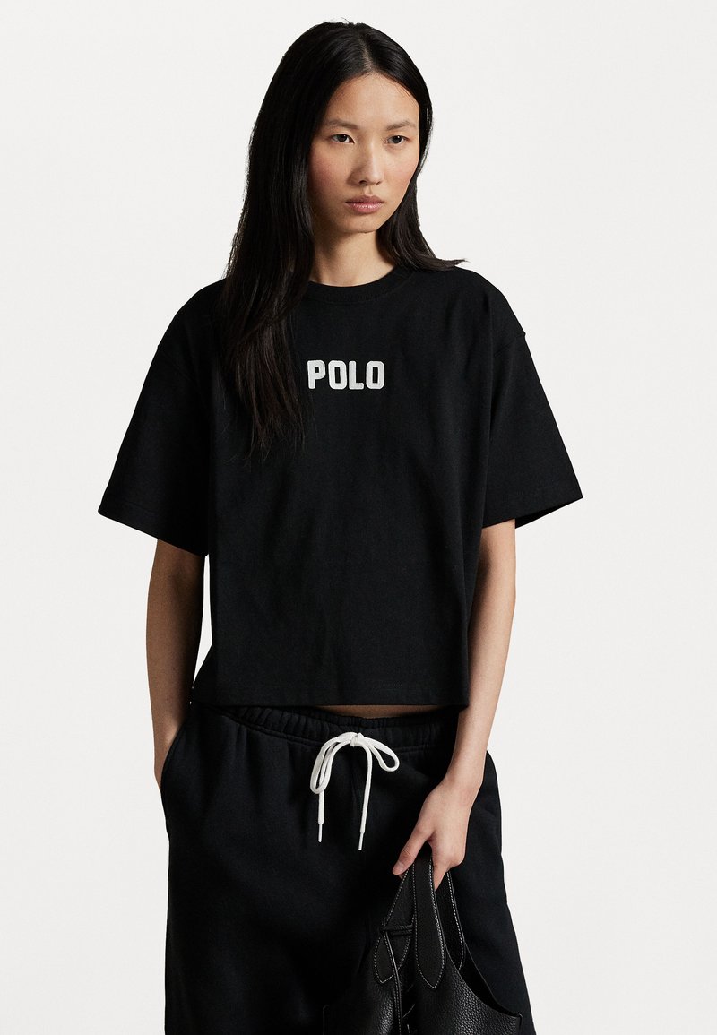 Tricou scurt negru din bumbac, cu "POLO" impricrat în alb. Asortat cu pantaloni de trening negri, largi, și o geantă neagră cu șnur.