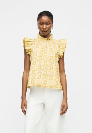 Femme portant un chemisier sans manches jaune à volants fleuris associé à un pantalon blanc à jambes larges sur un fond uni.