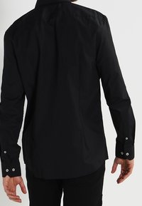 Camisetta nera a maniche lunghe con colletto classico, polsini abbottonati e una texture di tessuto liscia. Vestibilità tailored con orlo curvo.