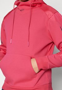 Różowa bluza z kapturem, z przednią kieszenią, charakteryzująca się gładkim materiałem i ściągaczami przy mankietach. Logo nadrukowane na piersi, sznurki przy kapturze.