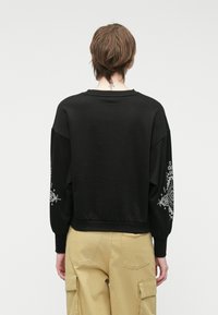 Melnā kapuce ar pārmērīgu piegriezumu, ribota apkakle un dekoratīvs balts prints uz piedurknēm. Materiāls ir gluds ar vieglu elastību.