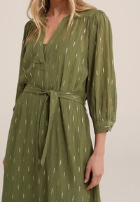 Mörkgrön, långärmad klänning med V-hals och framskjort placket. Har en knytbar midjebälte och gul tråddekoration i ett strukturerat mönster.