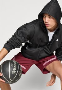 Sweat à capuche noir zippé avec capuche, arborant un logo blanc et des poches latérales. Shorts rouges avec des accents roses. Tenant un ballon de basket noir.