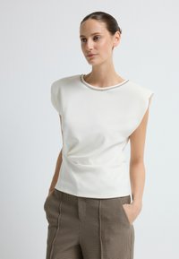 Biała bluzka z krótkim rękawem o marszczonym fasonie i dekoracyjnym srebrnym wykończeniu wokół dekoltu, zestawiona z brązowymi spodniami w kratę.