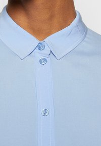 Lys blå skjorte med knapper, der har en krave, tre knapper på knæstykke og et glat, let struktureret stof.