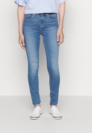 Persoon draagt lichtblauwe skinny jeans, een lichtblauw shirt en witte sneakers, staand tegen een effen achtergrond.