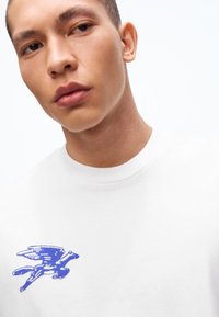 Fiatal férfi fehér ingben, melynek mellkasán kék szárnyas ló grafikája látható, kissé oldalra néz, fehér háttér előtt.