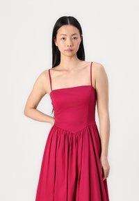 Vestido fucsia con tirantes finos, escote cuadrado y falda recogida. Hecho de una tela suave, con un torso ajustado y un fondo acampanado.