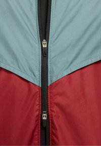 Väriestäävä takki, jossa on vaaleansininen yläosa ja punainen alaosa. Edessä vetoketju mustilla yksityiskohtilla ja sileä synteettinen pinta.