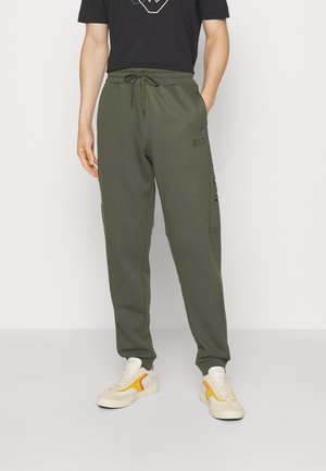 Mężczyzna ubrany w oliwkowozielone spodnie dresowe z bocznymi kieszeniami oraz białe trampki z żółtymi i pomarańczowymi akcentami, stojący na białym tle.