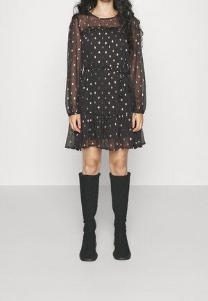Robe noire avec manches longues transparentes, motif à pois, corsage ajusté, jupe à volants superposés. Associée à des bottes en daim noires montant jusqu'aux genoux.