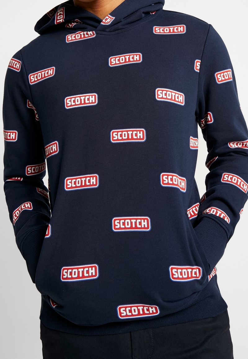 Sudadera marina hecha de un tejido suave con un patrón de logotipos repetidos en rojo y blanco. Cuenta con capucha con cordón y puños y dobladillo acanalados.
