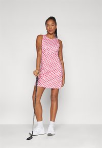 Biała i czerwona sukienka golfowa w wzory, bez rękawów. Modelka trzyma kij golfowy, ubrana w białe trampki i skarpetki do kostek.