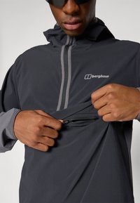 Čierna ľahká bunda s šedými akcentmi a zipsovou kapsou, obsahujúca kapucňu a predný polovičný zips. Logá viditeľné na povrchu látky.