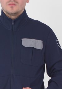 Giacca zip-up blu navy con colletto alto e tasca grigia sul lato sinistro del petto. Tessuto liscio con un design aderente, adatta per un look casual.