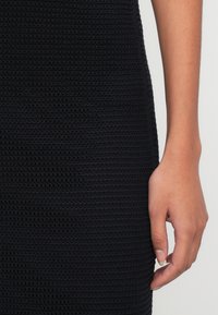 Rochie neagră tricotată cu un model texturat în rib, cu o primăvară a unei mâini care apucă țesătura. Material moale și structurat.