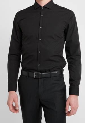 Mann trägt ein eng anliegendes schwarzes Langarmhemd mit Knöpfen, in schwarze Hose gesteckt, mit einem schwarzen Ledergürtel vor einem einfarbigen Hintergrund.