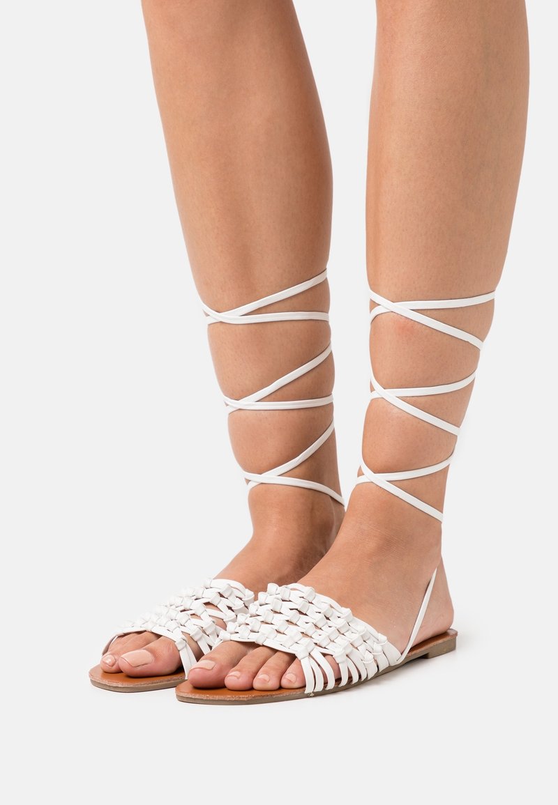 Jambes nues portant des sandales blanches plates avec des brides tissées et de longues fines lanières enroulées autour des mollets sur un fond clair.