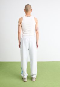 White sleeveless tank top and light grey sweatpants with blue side stripes; model stands with a plain white backdrop and green flooring.
