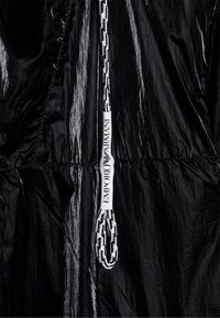 Svart pufferjacka med glänsande, krinklad textur; har en tjock vit och svart dragsko med "EMPORIO ARMANI"-logotyp och en slät yta.