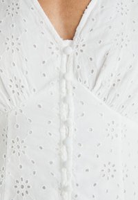 Biele bavlnené šaty s vyšívanými kvetinovými vzormi, s výstrihom do V a dekoračnými gombíkmi pozdĺž prednej časti, s mierne textúrovanou tkaninou.