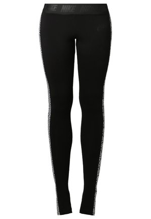 Femme debout pieds nus portant un t-shirt blanc à manches courtes et col rond, et un legging noir avec des bandes latérales blanches à motifs.