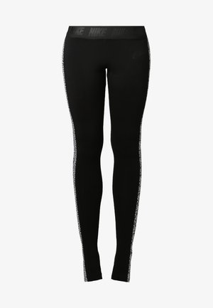 Vrouw staat blootsvoets, gekleed in een wit shirt met korte mouwen en ronde hals en zwarte legging met witte patroonstrepen aan de zijkant.