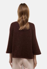 Femme aux cheveux ondulés mi-longs portant un pull en peluche marron foncé et un pantalon beige clair, debout de dos sur un fond uni.