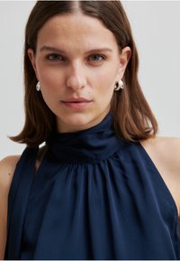 Marineblauw haltertopje gemaakt van gladde stof met plooien rondom de halslijn. Het model draagt zilveren oorringen met een minimalistisch ontwerp.