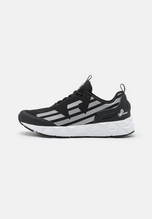 Sneaker atletica nera e grigia con suola geometrica bianca, etichette per il sollevamento, lacci e dettagli del logo laterale su sfondo bianco semplice.
