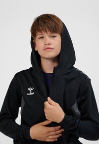 Schwarzer Kapuzenpullover mit strukturierter Stoffoberfläche, Reißverschluss vorne und Logo auf der Brust. Mit grauen Akzenten an Schultern und Ärmeln.