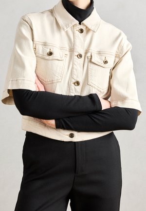 Person iført en beige cropped jakke over en sort langærmet top og sorte bukser med armene krydset ved taljen.