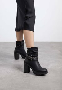 Botas de tobillo de cuero negro con puño caído, detalle de hebilla, acentos de tachuelas y tacón grueso. Suela texturizada para mayor tracción.