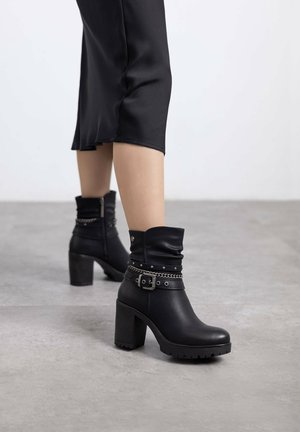 Botas de tobillo de cuero negro con puño caído, detalle de hebilla, acentos de tachuelas y tacón grueso. Suela texturizada para mayor tracción.